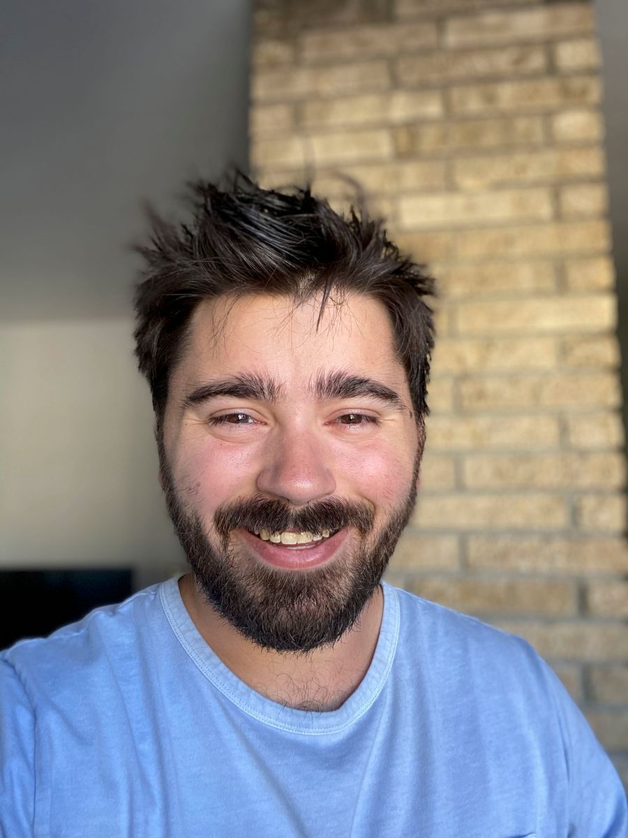 Eliot Fowler smiling in a light blue t-shirt, in front of a brick wall
